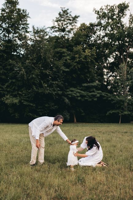 A man and a woman are outdoors in a grassy area, engaging with a young girl who is standing between them. The man, dressed in light-colored trousers and a white shirt, is bending slightly forward, hol
