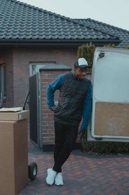 A man wearing a white polo shirt, black cap, and jeans stands inside the open back of a commercial van, which is filled with numerous packed cardboard boxes of various sizes. The boxes are arranged ne