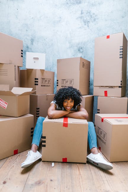 A young man with long hair, wearing a blue jumpsuit, a headband, and red running shoes, is sitting on a wooden floor surrounded by several large cardboard boxes sealed with red and black tape. The boxes are stacked and arranged around him inside a bright room with white walls and large arched windows that let in natural daylight. Outside the windows, a residential building and trees are visible. The man has a relaxed expression, with his arms resting on his knees, and appears to be taking a break during a home relocation or furniture transport process. Behind him, there are additional boxes and packaging materials, suggesting an ongoing packing and moving project. This scene, captured by Man and Van Wallington, illustrates the preparation phase of a house removal, focusing on packaging, space organization, and moving logistics within a residential setting.
