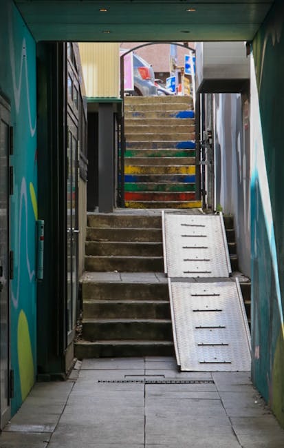 A top-down view of a narrow staircase with light-colored, possibly stone or concrete steps, enclosed by metal railings and walls. The central metal handrail runs along the middle of the stairs, design