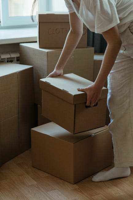 A person wearing a white t-shirt and blue jeans is kneeling on a concrete floor inside a property, using a red marker to write on a brown cardboard box. The box is secured with white packing tape acro