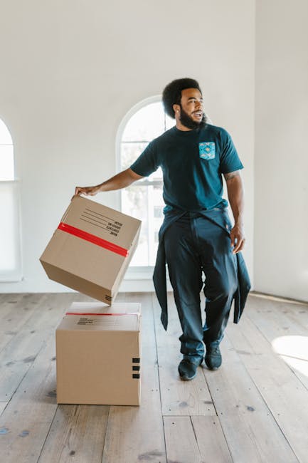 Two individuals involved in a house relocation process, sorting and handling cardboard boxes of various sizes within a room. The man on the left, wearing a white t-shirt and blue jeans, is lifting a m