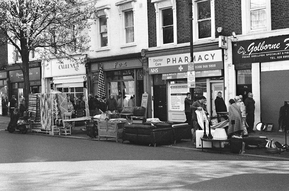 A black and white photograph of a busy street scene on Wallington High Street, with various items of furniture, including sofas, cardboard boxes, and wrapped household goods, arranged outside a row of